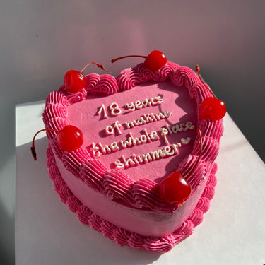 Розова торта във форма на сърце с черешки, кремав кант и надпис '18 years of making the whole place shimmer ♡'