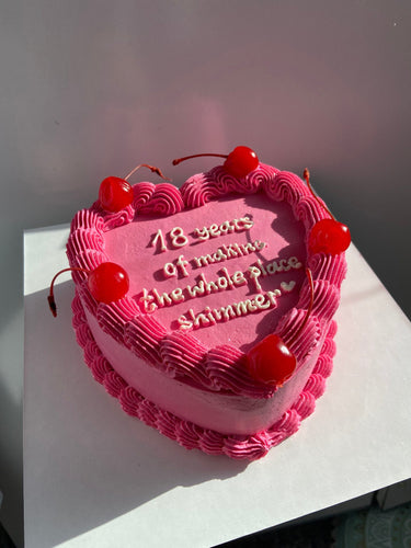 Розова торта във форма на сърце с черешки, кремав кант и надпис '18 years of making the whole place shimmer ♡'