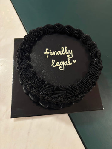 Кръгла черна торта с кремав кант и надпис 'finally legal♡' на горната повърхност.