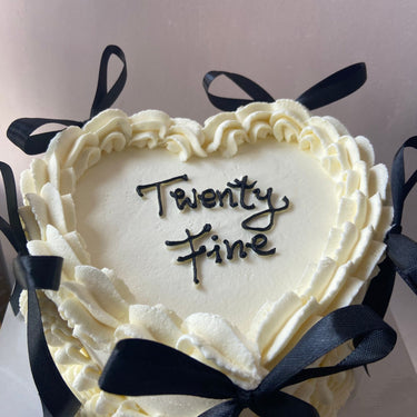 White heart-shaped cake with decorative ribbons, a cream border and the message "twenty fine".