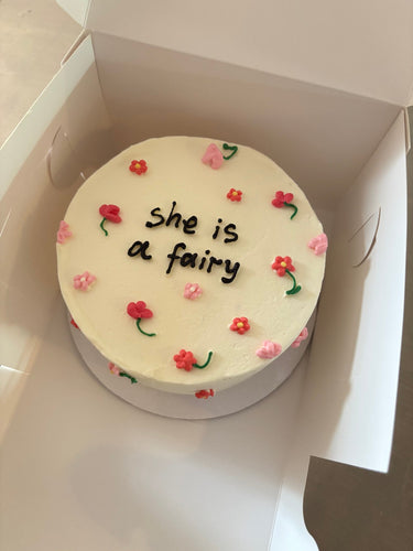 Кръгла бяла торта с рисунка на фея и надпис "she is a fairy" върху глазурата.