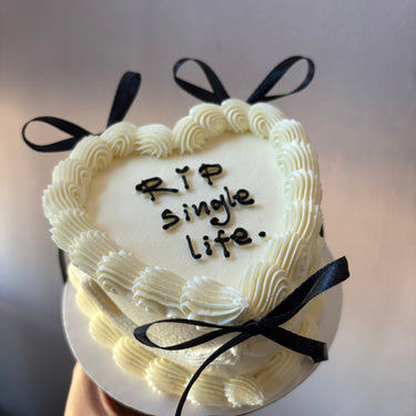 Cream heart cake with the inscription RIP single life, decorated with ribbons and a cream border.