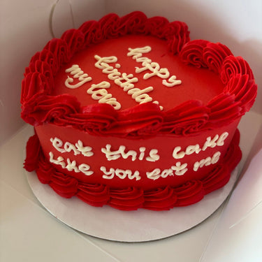 Червена торта във форма на сърце с кремав кант и страничен надпис 'happy birthday, my love / eat this cake like you eat me'.