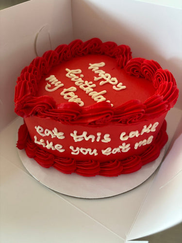 Червена торта във форма на сърце с кремав кант и страничен надпис 'happy birthday, my love / eat this cake like you eat me'.