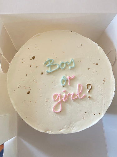 Кръгла бяла торта с надпис 'Boy or girl?' в златни букви.