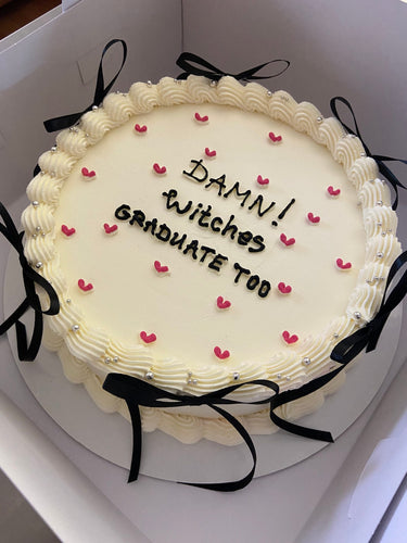 Кръгла кремава торта с надпис DAMN! Witches GRADUATE TOO, украсена с перли, панделки и кремав кант.