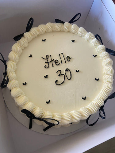 Кръгла бяла торта с надпис Hello 30, кремав кант и декоративни панделки.