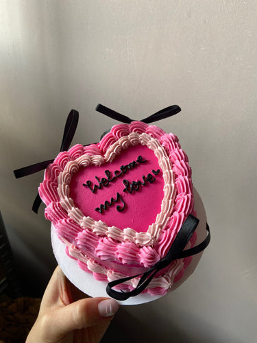 Розова торта във форма на сърце с надпис Welcome my love, кремав кант и декоративни панделки.