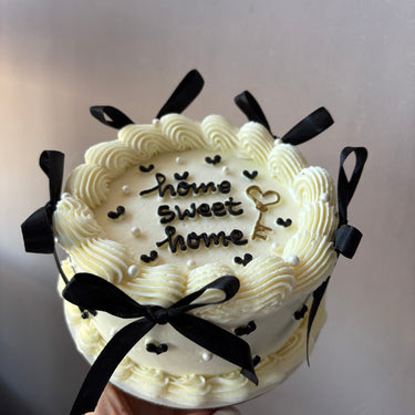 A round cream-coloured cake with the inscription 'home sweet home', decorated with pearls, ribbons and a cream border.