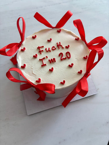 Кръгла бяла торта с надпис "Fuck I'm 20" и декоративни панделки.