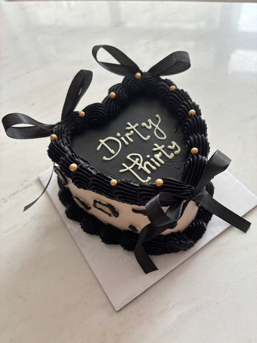 Черна торта във формата на сърце с надпис "Dirty Thirty", украсена с панделки, перли, кремав кант и рисунка.