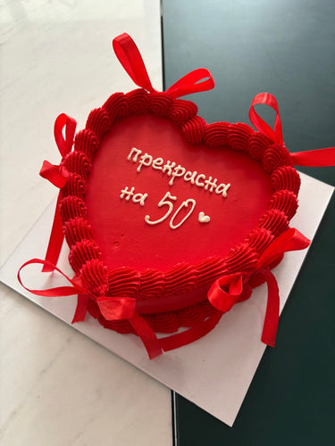 Кръгла червена торта с надпис "прекрасна на 50", кремав кант и декоративни панделки.