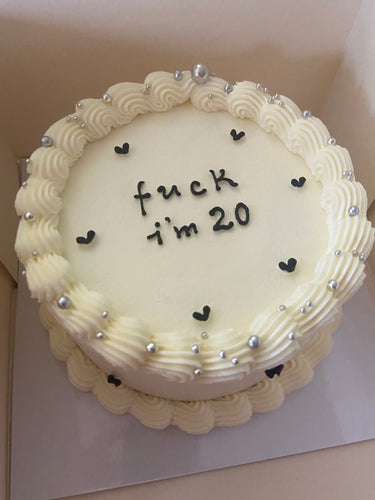 Кръгла кремава торта с надпис 'fuck i'm 20', украсена с перлени декорации и кремав кант.