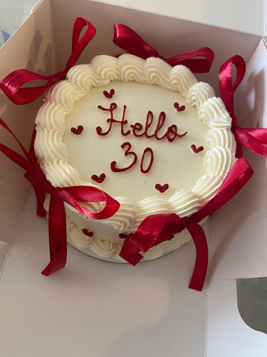 Кръгла кремава торта с надпис 'Hello 30', декорирана с панделки и кремав кант по ръба.