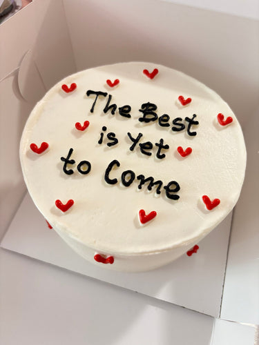 Кръгла бяла торта с надпис 'The Best is Yet to Come' върху глазурата, без допълнителна украса.