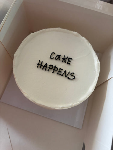 Кръгла бяла торта с надпис CAKE HAPPENS в тъмни букви, без допълнителна украса.