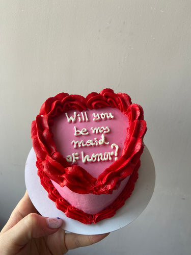 Розова торта във форма на сърце с кремав кант и надпис "Will you be my maid of honor?"