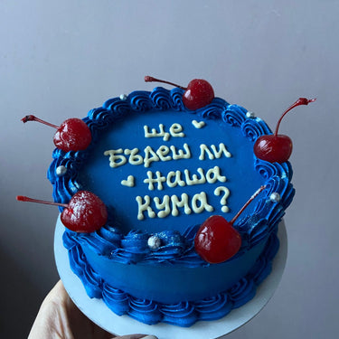 Кръгла синя торта с надпис 'Ще бъдеш ли наша кума?', украсена с черешки, бели перли и кремав кант.