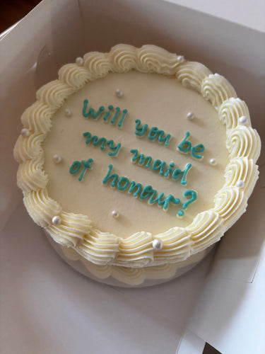 Кръгла кремава торта с надпис 'Will you be my maid of honour?', украсена с перли и кремав кант.