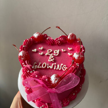 Розова торта във формата на сърце с надпис '29 & GLOWING', украсена с черешки, панделки, кремав кант, перли и брокат.