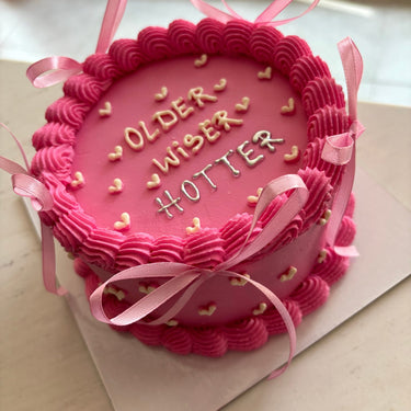Кръгла розова торта с кремав кант и панделки, с надпис OLDER WISER HOTTER