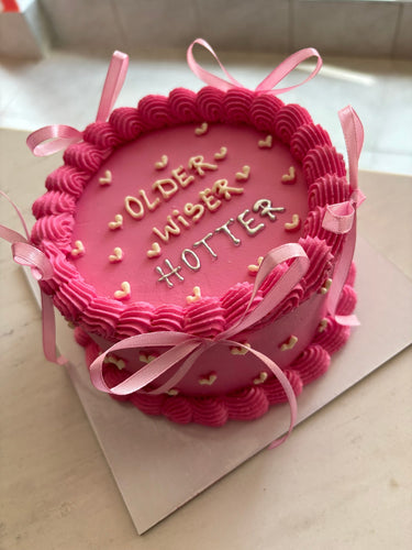 Кръгла розова торта с кремав кант и панделки, с надпис OLDER WISER HOTTER