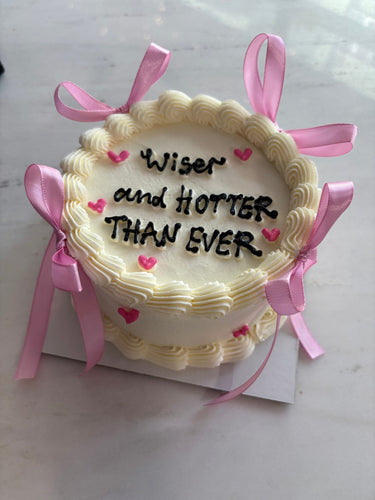 Кръгла кремава торта с надпис 'Wiser and HOTTER THAN EVER', декорирана с панделки и кремав кант.