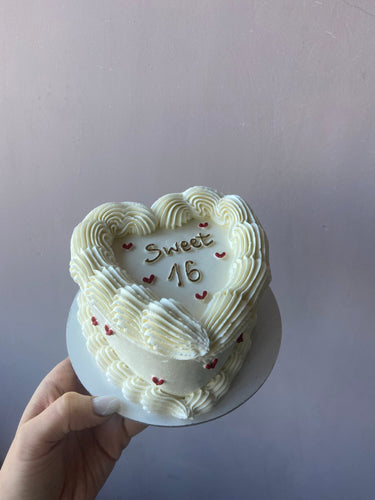 Кремава торта във форма на сърце с надпис Sweet 16 в злато и кремав декоративен кант по краищата.