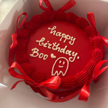 Кръгла червена торта с кремав кант, рисунка и панделки, надпис 'happy birthday, Boo' с сърце.