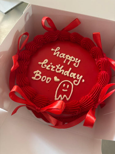 Кръгла червена торта с кремав кант, рисунка и панделки, надпис 'happy birthday, Boo' с сърце.