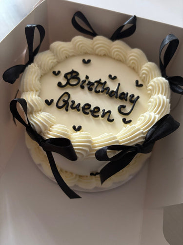 Кръгла бяла торта с надпис Birthday Queen, украсена с панделки и кремав кант.