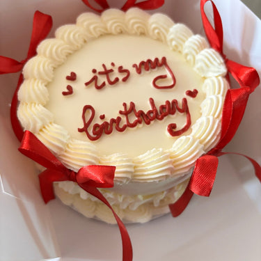 Кръгла кремава торта с надпис 'it's my birthday', украсена с панделки и кремав кант.