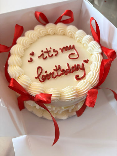 Кръгла кремава торта с надпис 'it's my birthday', украсена с панделки и кремав кант.