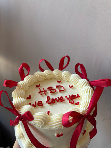 Кръгла бяла торта с панделки, сърца и кремав кант, с надпис HBD we love you