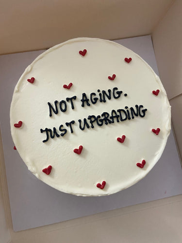Кръгла бяла торта с надпис NOT AGING. JUST UPGRADING в тъмни букви върху гладка бяла глазура.