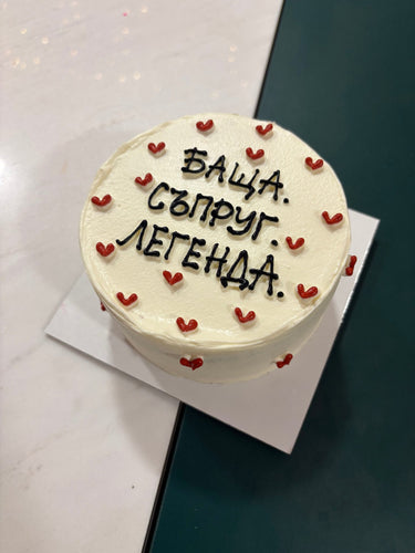 Кръгла бяла торта с надпис БАЩА. СЪПРУГ. ЛЕГЕНДА. в черни букви върху гладка глазура.