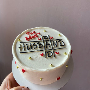 Кръгла бяла торта с надпис "The best HUSBAND AND DAD", украсена с перли и рисунка.