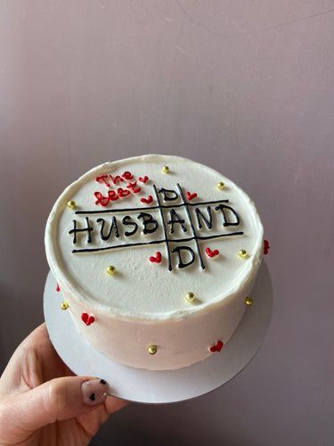 Кръгла бяла торта с надпис "The best HUSBAND AND DAD", украсена с перли и рисунка.