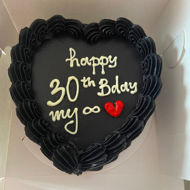A black heart-shaped cake with a cream border and the inscription 'happy 30th Bday my ∞ ♥'
