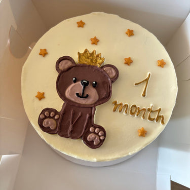 A round cream cake with an illustration of a bear with a crown and a gold inscription for a baby's month-birthday.