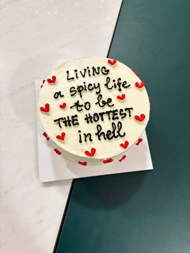 Бяла кръгла торта с надпис 'Living a spicy life to be THE HOTTEST in hell' върху глазурата, без допълнителна украса.
