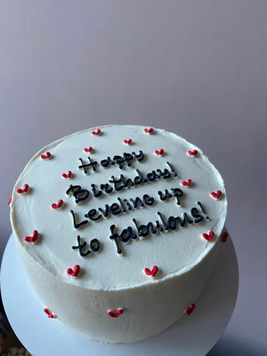 Бяла кръгла торта с надпис 'Happy Birthday! Leveling up to fabulous!' върху глазурата, без допълнителна украса.
