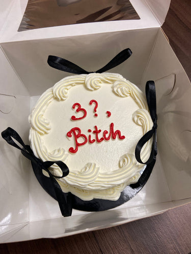 Кръгла бяла торта с кремав кант и панделки, надпис '3?, Bitch' отгоре.