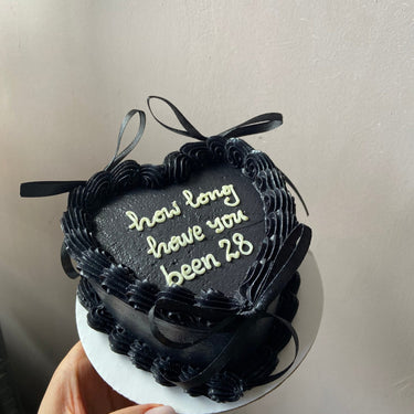 Heart-shaped cake with black icing, a cream border, ribbons and the inscription 'how long have you been 28' on top.