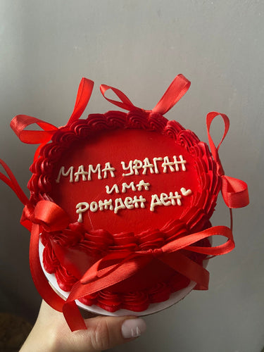 Кръгла червена торта с кремав кант, панделки и надпис 'МАМА УРАГАН има рожден ден' отгоре.
