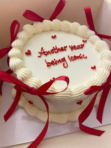 Кръгла бяла торта с кремав кант, панделки и надпис 'Another year being iconic' отгоре.