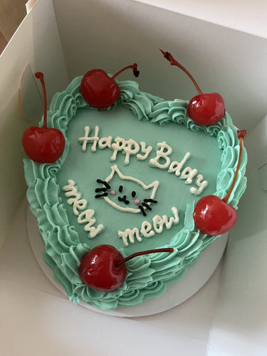Сърцевидна ментова торта с рисунка на коте, черешки, кремав кант и надпис 'Happy Bday meow meow meow'.