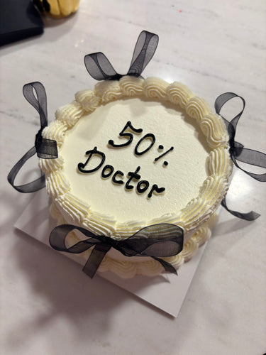 Кръгла бяла торта с панделки, кремав кант и надпис '50% Doctor'.