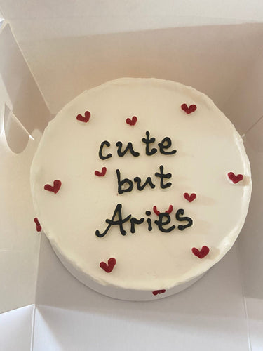 Кръгла бяла торта с надпис 'cute but Aries' на повърхността, без допълнителна украса.