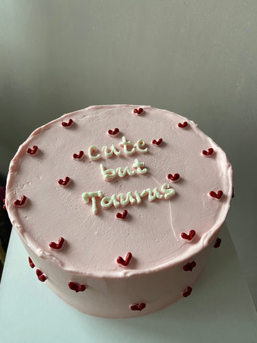 Кръгла бебешко розова торта с надпис 'cute but Taurus' на повърхността, без допълнителна украса.